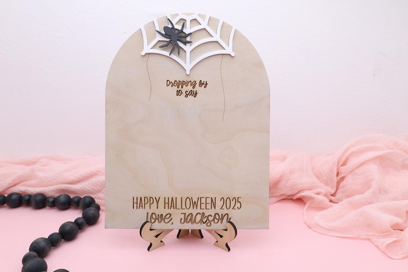 May include: A wooden Halloween sign with a spider web and spider design. The sign reads "Dropping by to say" and "Happy Halloween 2025 Love, Jackson". The sign is supported by a wooden stand.