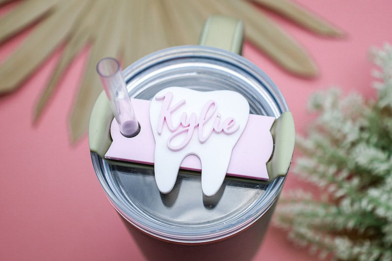 May include: A pink and white tooth-shaped tumbler charm with the name "Kylie" written in pink cursive letters. The charm is attached to a pink and green tumbler lid.