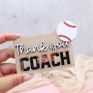 May include: A wooden baseball-themed gift with the words "Thank you COACH" cut out. The word "COACH" is in black block letters with orange accents. A small, round baseball with red stitching is attached.