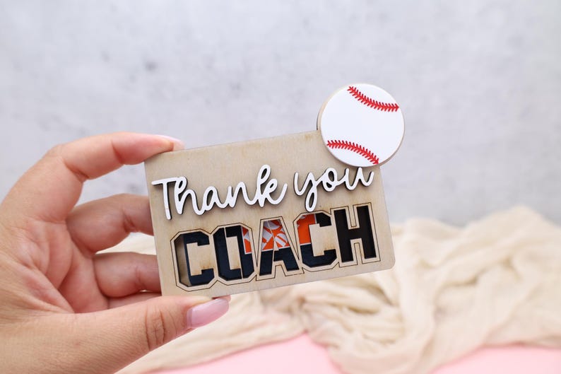 May include: A wooden baseball-themed gift with the words "Thank you COACH" cut out. The word "COACH" is in black block letters with orange accents. A small, round baseball with red stitching is attached.