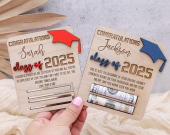 May include: Two wooden graduation cards with a graduation cap design. The cards are personalized with the names "Sarah" and "Jackson" and the year "2025". The card for Sarah has a red graduation cap and the card for Jackson has a blue graduation cap. Both cards have a slot for money.