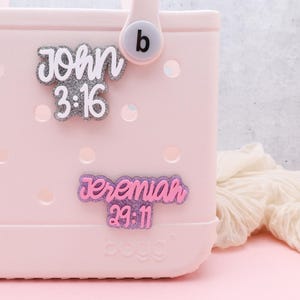 May include: Pink and white tote bag with two personalized charms. The top charm is silver glitter with the text "John 3:16". The bottom charm is pink and purple glitter with the text "Jeremiah 29:11".