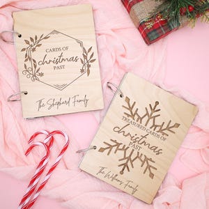 Puede incluir: Dos álbumes de fotos de madera con encuadernación de anillas. El álbum superior tiene un diseño floral y el texto "CARDS OF christmas PAST The Shepherd Family". El álbum inferior tiene un diseño de copo de nieve y el texto "TREASURED CARDS OF christmas PAST The Williams Family".