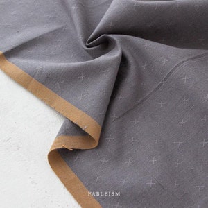 May include: Close-up of a gray fabric with a cross-stitch pattern. The fabric has a tan border and is folded, showing texture and detail. The word "FABLEISM" is printed at the bottom.