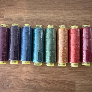 May include: An overhead shot displays eight spools of thread in various colors, including purple, blue, green, orange, and red. Each spool has a yellow label at the base. The spools are arranged horizontally on a wooden surface.