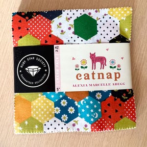 May include: A fabric charm pack with a variety of colorful hexagonal patterns. The pack is wrapped with a band featuring the text "catnap" and a pink tiger illustration. The pack is 5 inches in size and contains 42 pieces.