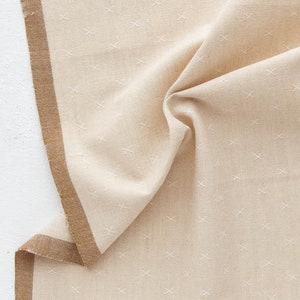 May include: Beige fabric with a repeating white cross pattern. A brown border runs along one edge. The fabric is folded, showing texture and dimension. Ideal for home decor or sewing projects.
