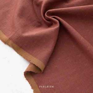 May include: A close-up of a rust-colored fabric with a subtle crosshatch pattern. The fabric has a soft, textured feel and a slightly sheen finish. The fabric is folded and draped over a white surface.