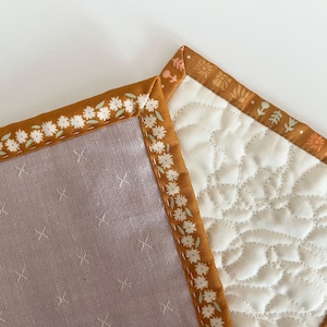 May include: Close-up of two quilted fabric squares. One is a light purple with white cross-stitch details. The other is off-white with a quilted pattern. Both have a brown floral border with white daisy accents.
