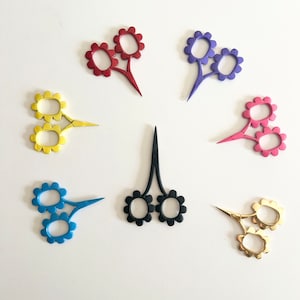 May include: Set of seven flower-shaped scissors in various colors, including red, purple, blue, black, and gold. The scissors have two flower-shaped blades and a single handle.