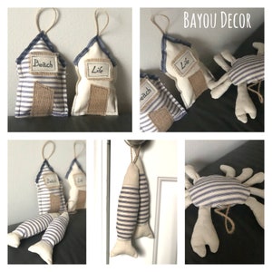 Puede incluir: Conjunto de adornos de tela hechos a mano con temática náutica. Los adornos incluyen una casa a rayas azules y blancas con la palabra "Beach" en ella, una casa a rayas azules y blancas con la palabra "Life" en ella, un pez a rayas azules y blancas y un cangrejo a rayas azules y blancas.