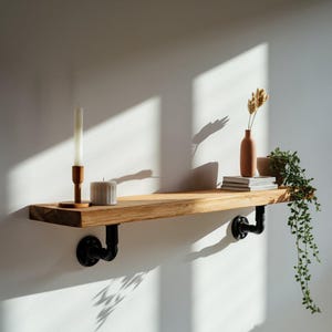 May include: A wooden shelf with black metal brackets, featuring a white candle, a small vase with dried flowers, and a stack of books. The shelf is mounted on a white wall with natural light streaming in.