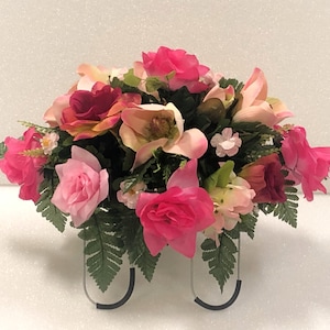 May include: A pink and white artificial flower arrangement with greenery, displayed on a silver stand. The arrangement features roses, lilies, and other flowers.