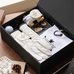 May include: A black gift box containing a white golf glove with the name Arthur, a white golf towel with a black golfer graphic, a white golf ball case with wooden tees, a golf ball, and a metal tool with the name Arthur printed on it.