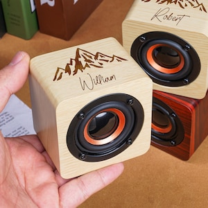 May include: A light-colored wooden cube speaker featuring a laser-engraved mountain graphic and the name William. The speaker has a black circular frame with an orange inner ring. A similar speaker with the name Robert is visible in the background.