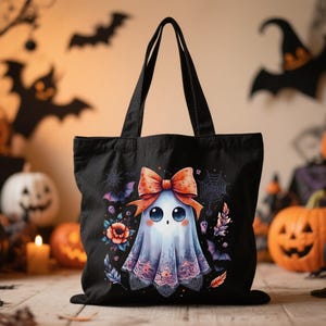 May include: A black tote bag featuring a central graphic of a small ghost with large eyes, wearing a large orange bow. The ghost is surrounded by colorful flowers, spider webs, and small bats. The bag has two black shoulder straps.