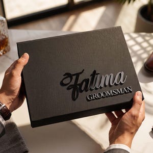 May include: A rectangular black gift box with a textured surface featuring the name Fatima in a large, raised script font and the word GROOMSMAN in a smaller, capitalised sans-serif font centred on the front lid of the box.