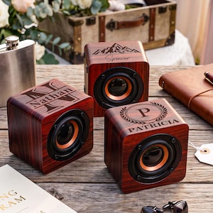 May include: Three cube-shaped wooden speakers with a dark wood grain finish. Each speaker features a black circular driver with an orange inner ring. The speakers are engraved with custom designs: one with a mountain graphic and the name Spencer, one with the name Nathan, and one with a laurel wreath, the letter P, and the name Patricia.