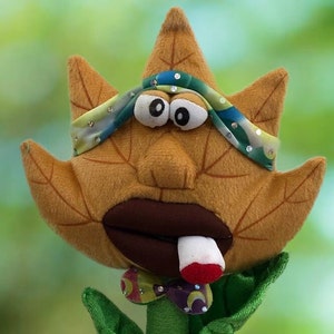 May include: A brown plush leaf puppet with a large mouth and googly eyes. The puppet is wearing a colorful headband and a bow tie. The puppet is holding a white cigarette in its mouth.