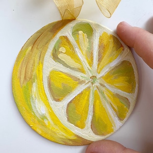 May include: A hand-painted round wooden ornament featuring a yellow lemon slice with a white center and green veins. The ornament is painted in a realistic style and has a glossy finish.