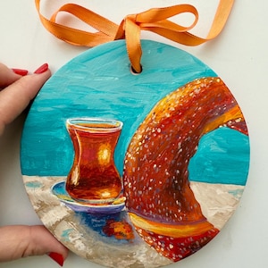 May include: A round wooden ornament painted with a glass of Turkish tea and a simit, a Turkish sesame seed bread, on a blue background. The ornament has a hole at the top for hanging.