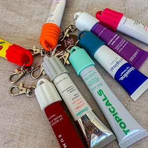 Puede incluir: Una colección de varios tubos de bálsamo labial, cada uno equipado con una tapa de silicona colorida y un llavero de mosquetón de metal. Los tubos presentan marcas como Glossier, Vaseline, Laneige y Topicals, con texto visible.