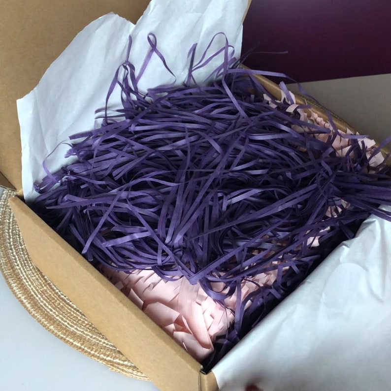 May include: A cardboard box filled with purple and pink shredded paper. The box is open and the shredded paper is visible.