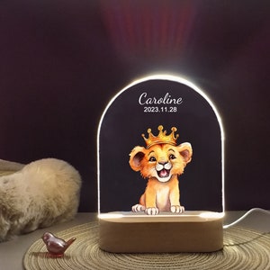 May include: A white and gold night light with a wooden base. The light features a cartoon lion cub wearing a gold crown. The text "Caroline 2023.11.28" is printed on the light.