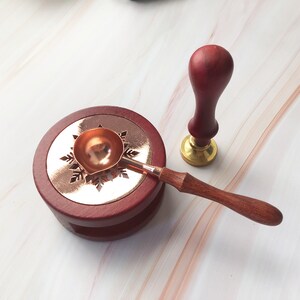 May include: A red wooden base with a snowflake design and a copper wax melting spoon with a wooden handle. A red wooden stamp with a gold base is also included.