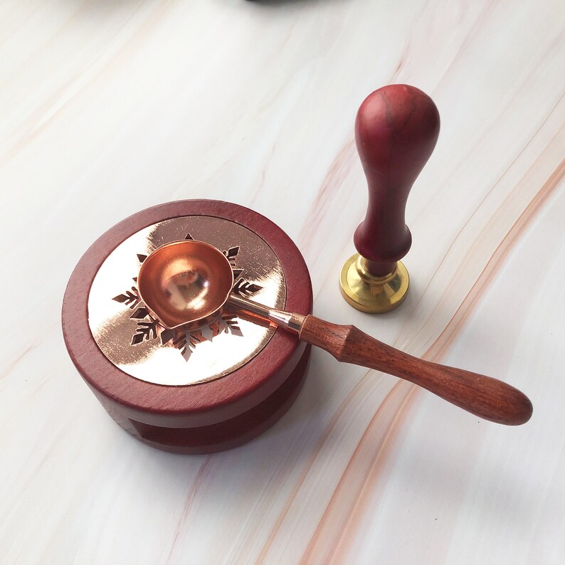 May include: A red wooden base with a snowflake design and a copper wax melting spoon with a wooden handle. A red wooden stamp with a gold base is also included.