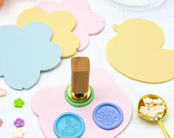 May include: A set of four pastel-colored wax seal stamps in the shape of a flower and a duck. The stamps are on a pink surface with three blue wax seals. A wooden handle for the stamp is also included.