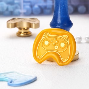 May include: An orange wax seal stamp with a cat-shaped controller design. A gold-colored stamp and a blue wax stick are also visible. The items are on a white surface, suggesting a crafting or stationery theme.