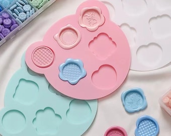 May include: Two silicone molds for making wax seals. The pink mold has six different shapes, including a butterfly, a circle, a cloud, a flower, a heart, and a square. The blue mold has five different shapes, including a circle, a cloud, a flower, a heart, and a square. There are also three wax seals in the image, one with a butterfly design, one with a flower design, and one with a square design.
