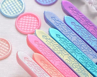 May include: A collection of multi-coloured wax seal sticks featuring an embossed filigree pattern, arranged alongside several small, round, grid-textured wax seals in shades of pink, blue, purple, and iridescent gold.
