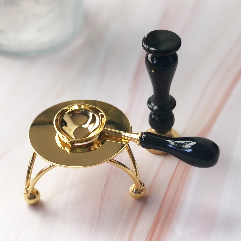 May include: A gold metal stand with a round top and three legs. A black and gold wax seal stamp with a wooden handle is on the stand.