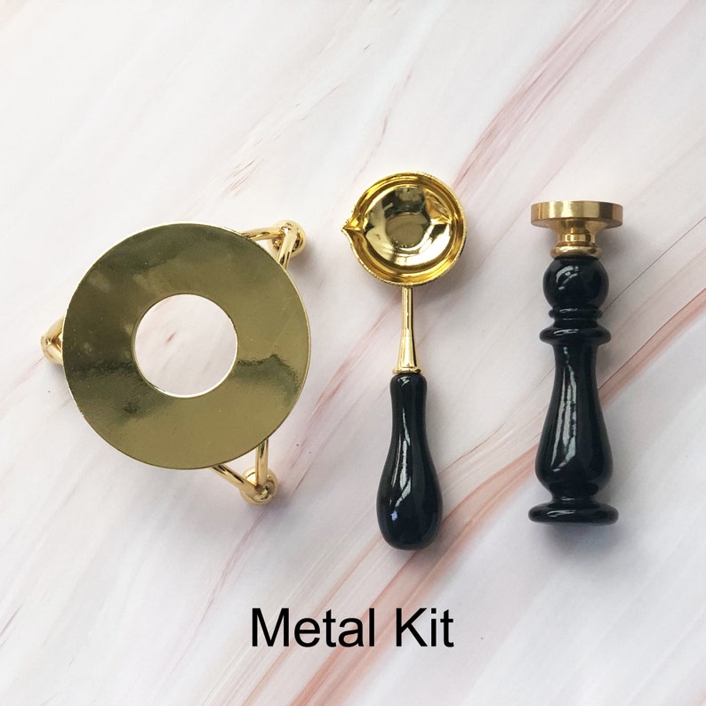 May include: A gold metal wax seal kit with a round stand, a spoon with a black handle, and a stamp with a black handle. The kit is on a white marble surface. Metal Kit.