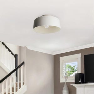 May include: A white, drum-shaped ceiling light fixture with a single exposed bulb. The light is mounted on a white ceiling in a room with a staircase and a window. The fixture has a simple, modern design.