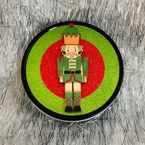 May include: A round, green and red glitter Christmas ornament with a wooden nutcracker figure in the center. The nutcracker is wearing a green and gold uniform with a red and white hat.