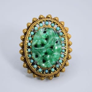 May include: An oval gold brooch with a carved jade centre depicting a floral design. The jade is encircled by small turquoise and pearl-like beads, and a decorative gold border. A vintage piece of jewellery.
