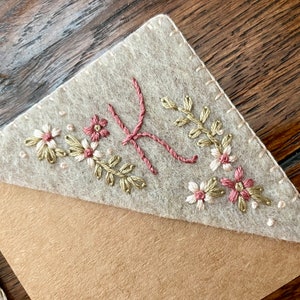 May include: A beige felt bookmark with a brown Kraft paper backing. The bookmark is decorated with embroidered flowers in shades of pink, white, and green.