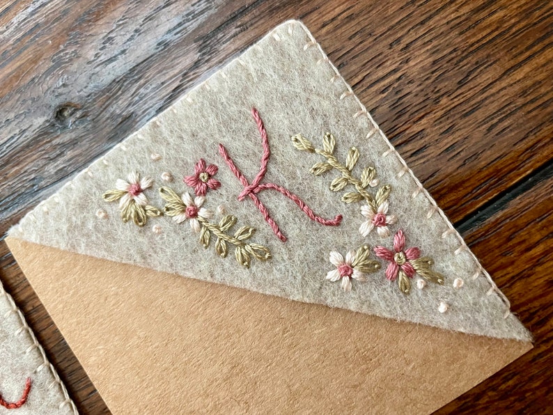 May include: A beige felt bookmark with a brown Kraft paper backing. The bookmark is decorated with embroidered flowers in shades of pink, white, and green.