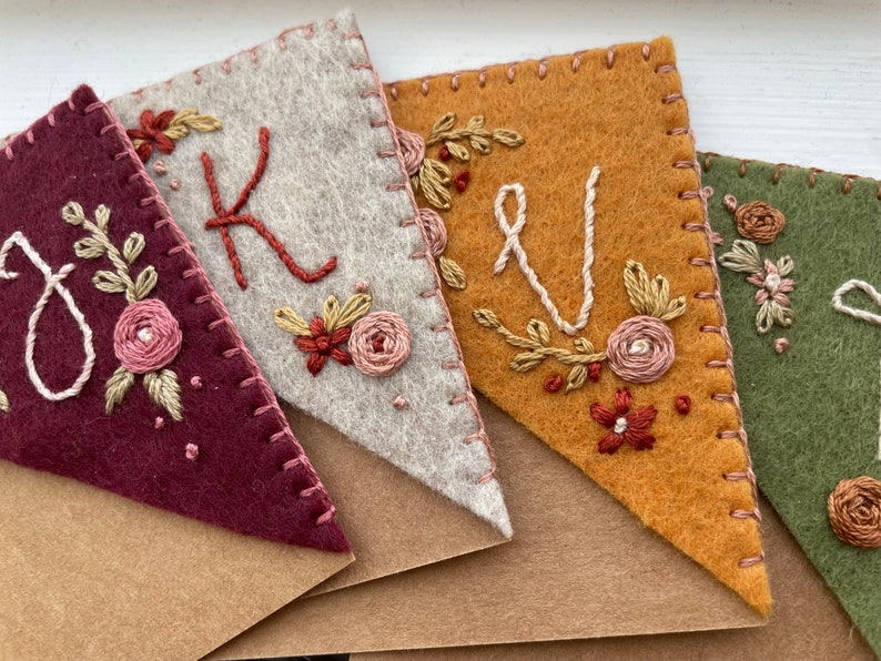 May include: A set of five felt pennants with embroidered floral designs and letters. The pennants are in shades of burgundy, gray, orange, green, and brown. The letters are K, V, and J.