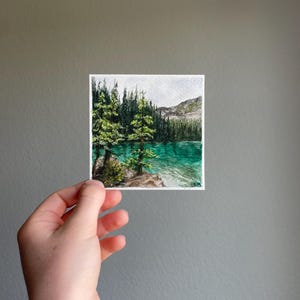 Peut inclure: Aquarelle repr&eacute;sentant un paysage lacustre serein. L'&oelig;uvre pr&eacute;sente un lac turquoise entour&eacute; d'arbres verts luxuriants et d'une cha&icirc;ne de montagnes en arri&egrave;re-plan. La peinture est tenue par une main sur un fond gris.