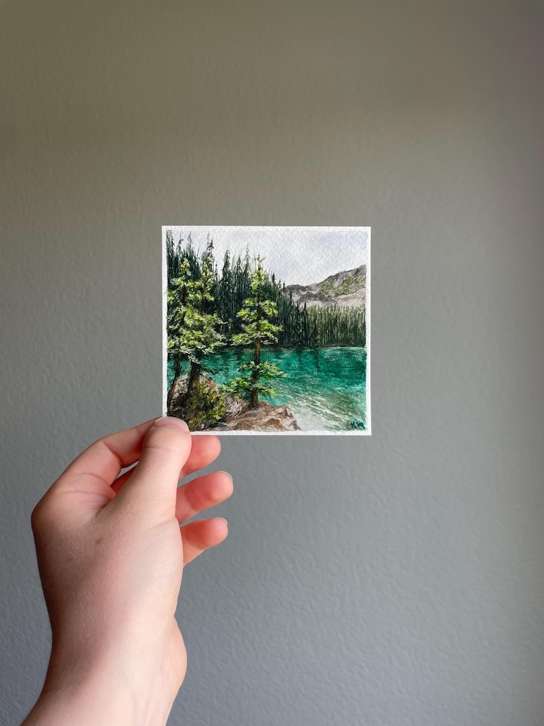 Peut inclure: Aquarelle repr&eacute;sentant un paysage lacustre serein. L'&oelig;uvre pr&eacute;sente un lac turquoise entour&eacute; d'arbres verts luxuriants et d'une cha&icirc;ne de montagnes en arri&egrave;re-plan. La peinture est tenue par une main sur un fond gris.