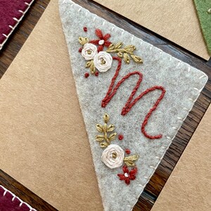 May include: A white felt triangle with a stitched border, featuring an embroidered capital letter "M" in red thread, surrounded by a floral design with white and gold flowers and red leaves.