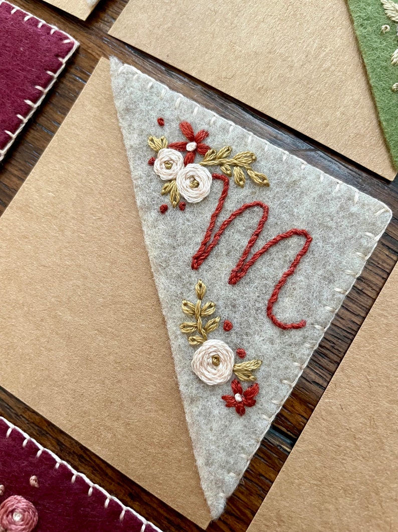 May include: A white felt triangle with a stitched border, featuring an embroidered capital letter "M" in red thread, surrounded by a floral design with white and gold flowers and red leaves.