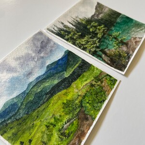 Peut inclure: Deux peintures &agrave; l'aquarelle de paysages de montagne. L'une repr&eacute;sente une cha&icirc;ne de montagnes vertes et bleues sous un ciel nuageux. L'autre repr&eacute;sente une montagne avec des arbres verts et un lac turquoise. Les deux sont sur papier blanc.