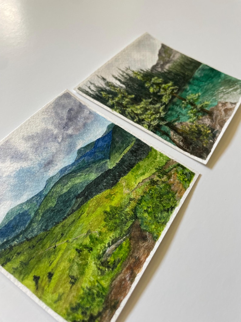 Peut inclure: Deux peintures &agrave; l'aquarelle de paysages de montagne. L'une repr&eacute;sente une cha&icirc;ne de montagnes vertes et bleues sous un ciel nuageux. L'autre repr&eacute;sente une montagne avec des arbres verts et un lac turquoise. Les deux sont sur papier blanc.
