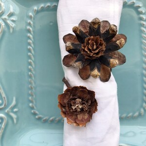 May include: A white napkin with two brown pine cone flower napkin rings. The napkin rings are on a light blue plate with a decorative border.