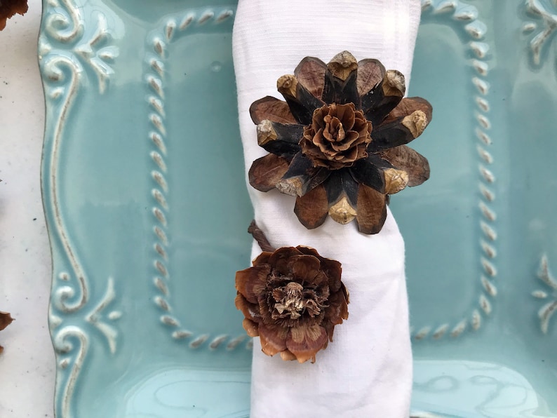 May include: A white napkin with two brown pine cone flower napkin rings. The napkin rings are on a light blue plate with a decorative border.